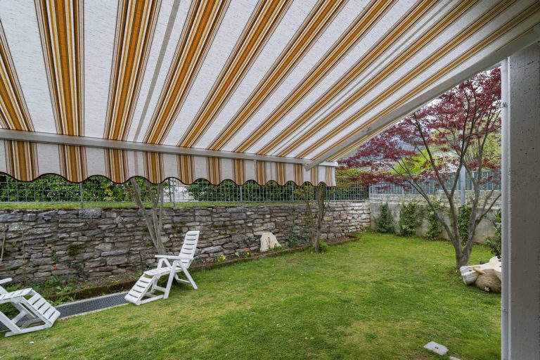 Tenda da sole a bracci aperta sul giardino, vista da sotto, con sdraio bianche sul prato